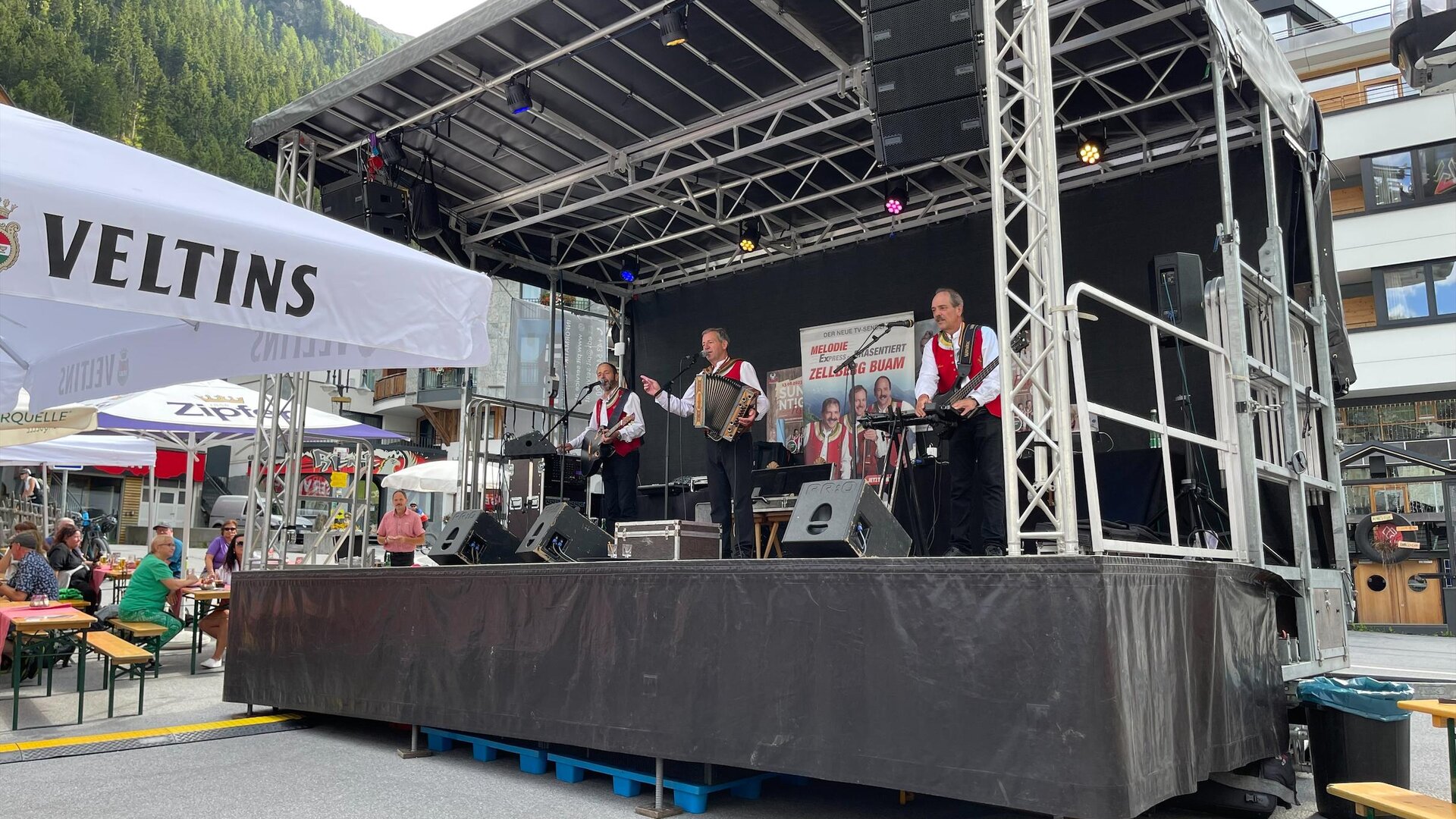 Eine Band spielt auf einer Bühne in einem Festivalambiente. Verweilte Gäste genießen die Musik unter einem klaren Himmel.