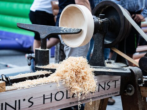 A woodworking machine processes wood into a bowl. Wood shavings are lying on the table and there are people visible in the background.