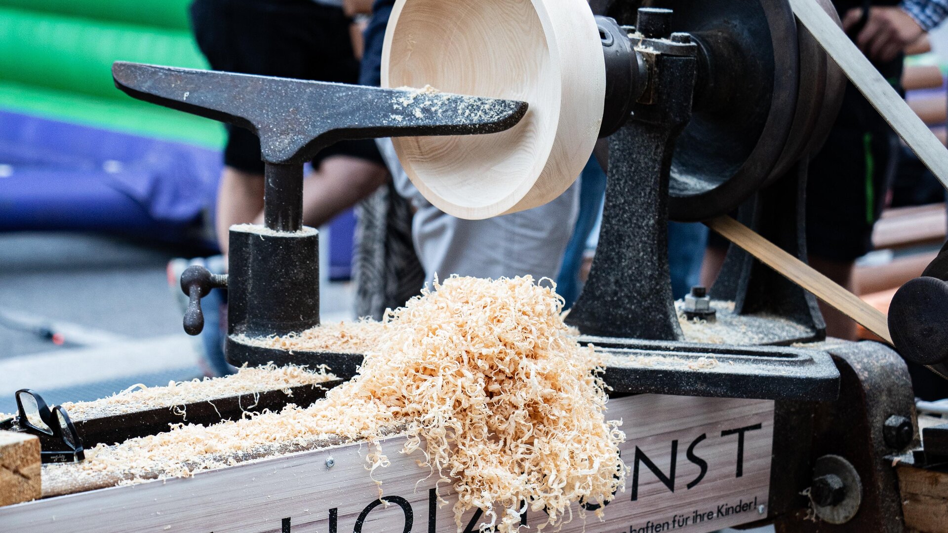 Eine Holzmaschine verarbeitet Holz zu einer Schüssel. Späne liegen auf dem Tisch und es sind Menschen im Hintergrund zu sehen.