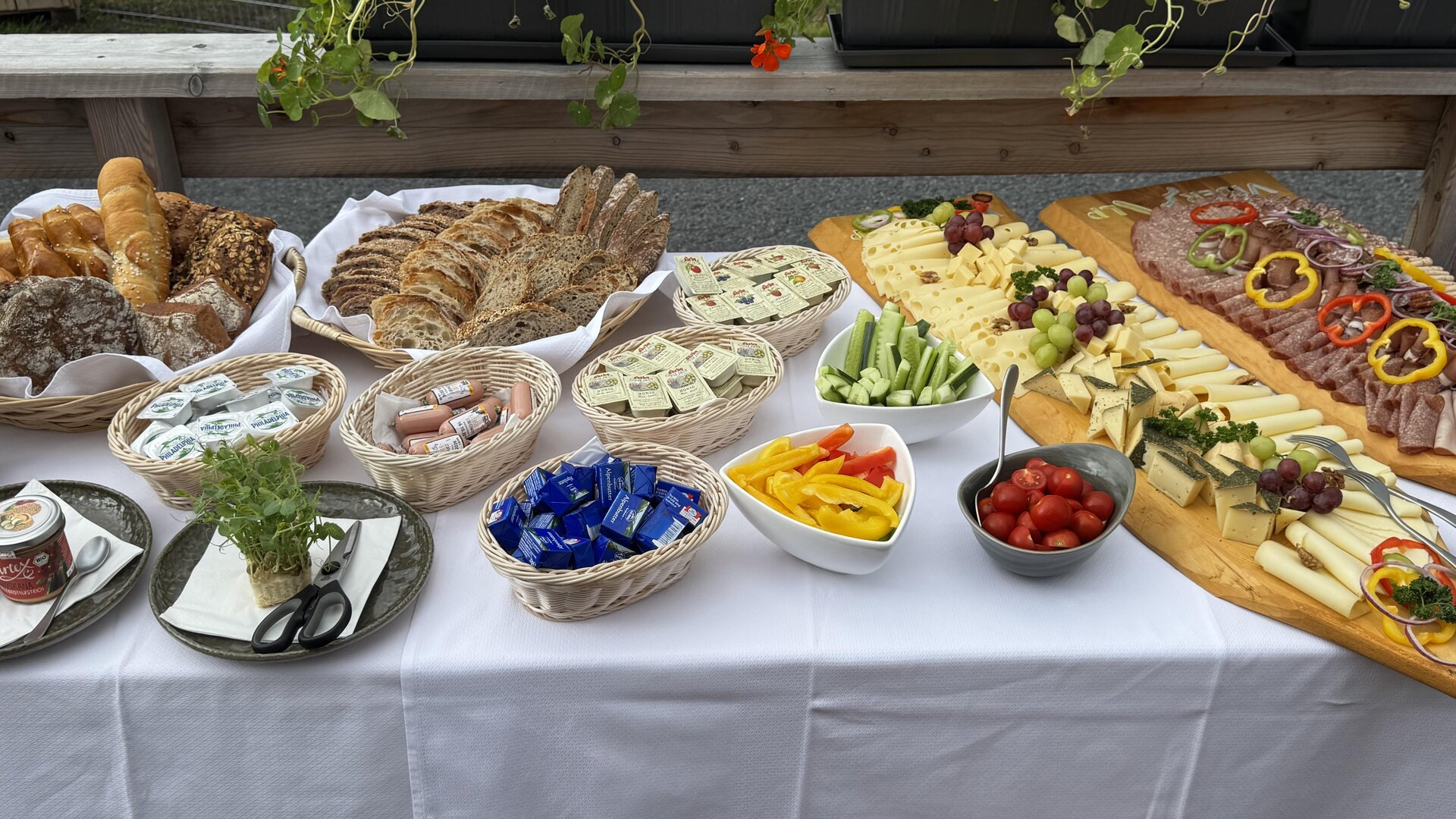 Ein reichhaltiges Buffet mit verschiedenen Brotsorten, Käse, Wurst und frischem Gemüse. Bunte Früchte und Snacks ergänzen das Angebot auf einem Tisch mit weißer Tischdecke.