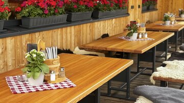 Ein gemütlicher Außenbereich mit Holztischen und bunten Blumen. Auf den Tischen stehen Gläser und frische Pflanzen, die eine einladende Atmosphäre schaffen.