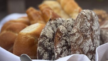 Ein Korb mit verschiedenen Brötchen und Brotsorten steht auf einem Tisch. Neben dem Brot befindet sich eine Schale mit Dip und einer essbaren Blume.