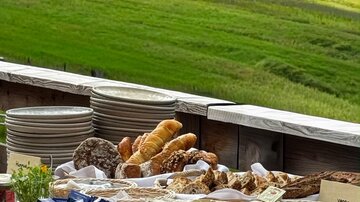 Ein schön angerichteter Tisch mit verschiedenen Speisen liegt vor einer herrlichen grünen Landschaft. Frisches Obst, Gemüse, Käse und Brot laden zum Genießen ein.