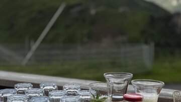 Ein Tisch mit Gläsern, Wasser und Saftflaschen steht vor einer schönen Berglandschaft. Die Atmosphäre wirkt entspannt und einladend.