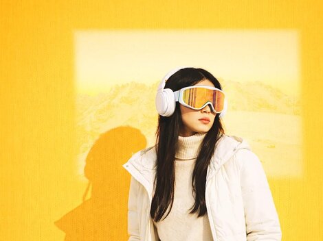 A woman stands in front of a yellow wall wearing a white jacket and colorful ski goggles. In the background, a blurred landscape is visible.