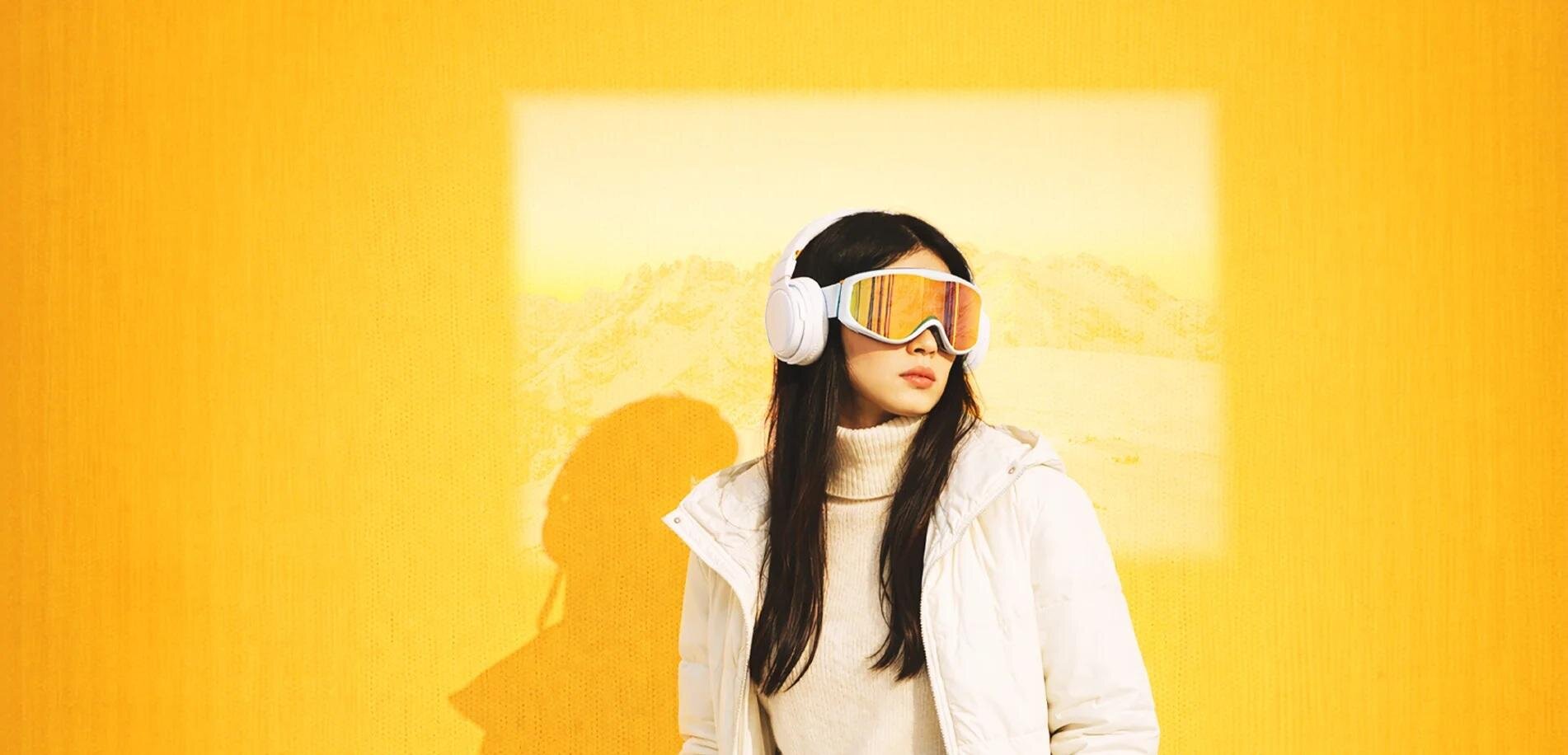 A woman stands in front of a yellow wall wearing a white jacket and colorful ski goggles. In the background, a blurred landscape is visible.