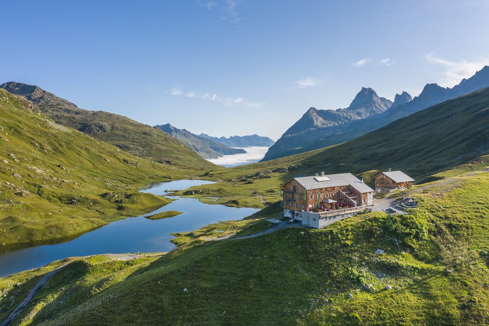 Ein modernes Holzgebäude liegt malerisch in den Bergen. Im Hintergrund sind schneebedeckte Gipfel und eine grüne Landschaft zu sehen.