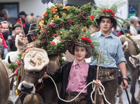 A festive procession with decorated cows and children in traditional attire. The animals wear colorful flower crowns and the background shows a large crowd of spectators.