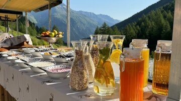 A beautifully set table outdoors with various drinks and dishes. In the background, mountains and a clear blue sky can be seen.