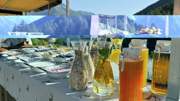 Ein schön dekorierter Tisch im Freien mit verschiedenen Getränken und Snacks. Im Hintergrund sind grüne Berge und ein klarer Himmel sichtbar.