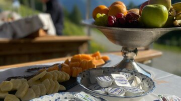Ein gedeckter Tisch mit verschiedenen Snacks, darunter Obst und Käse. Im Hintergrund sind Berge und eine schöne Landschaft zu sehen.