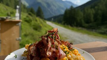 Ein Teller mit Rührei und knusprigem Speck, garniert mit Schnittlauch. Im Hintergrund erstrecken sich grüne Berge und ein blauer Himmel.