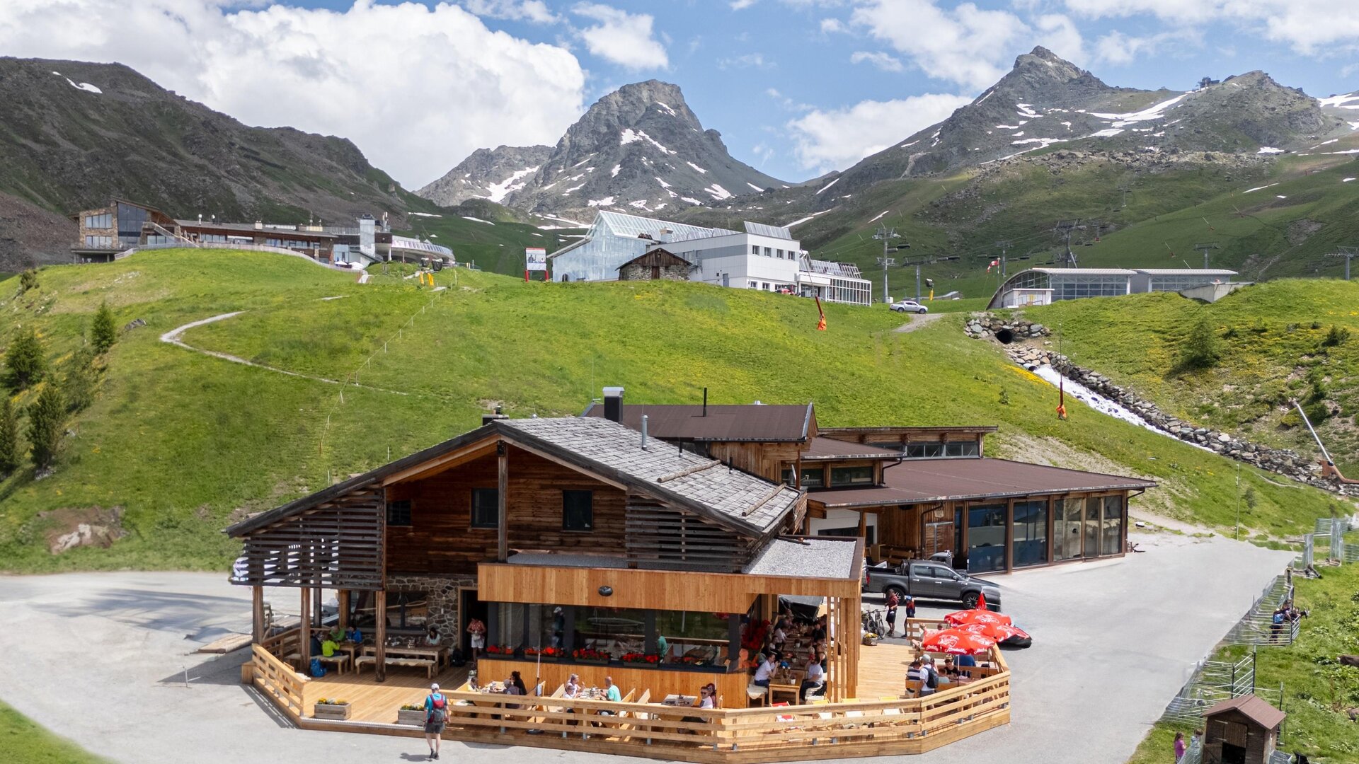 Eine rustikale Hütte in den Bergen mit Terrassenbereich. Im Hintergrund sind grüne Wiesen und schneebedeckte Gipfel zu sehen.