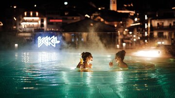 Two women enjoy the water in a lit pool at night. In the background, city lights are visible.