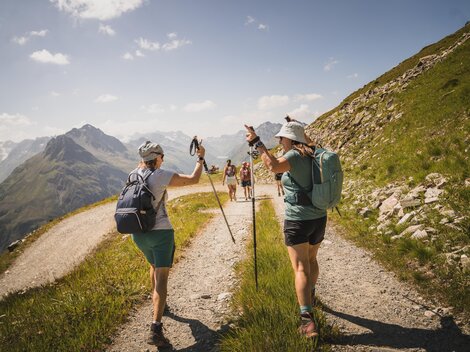 Two hikers on a mountain path enjoy nature. In the background, other hikers and mountains can be seen.