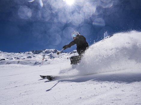A skier glides with energy through the fresh snow. In the background, there are snow-covered mountains and a clear blue sky.