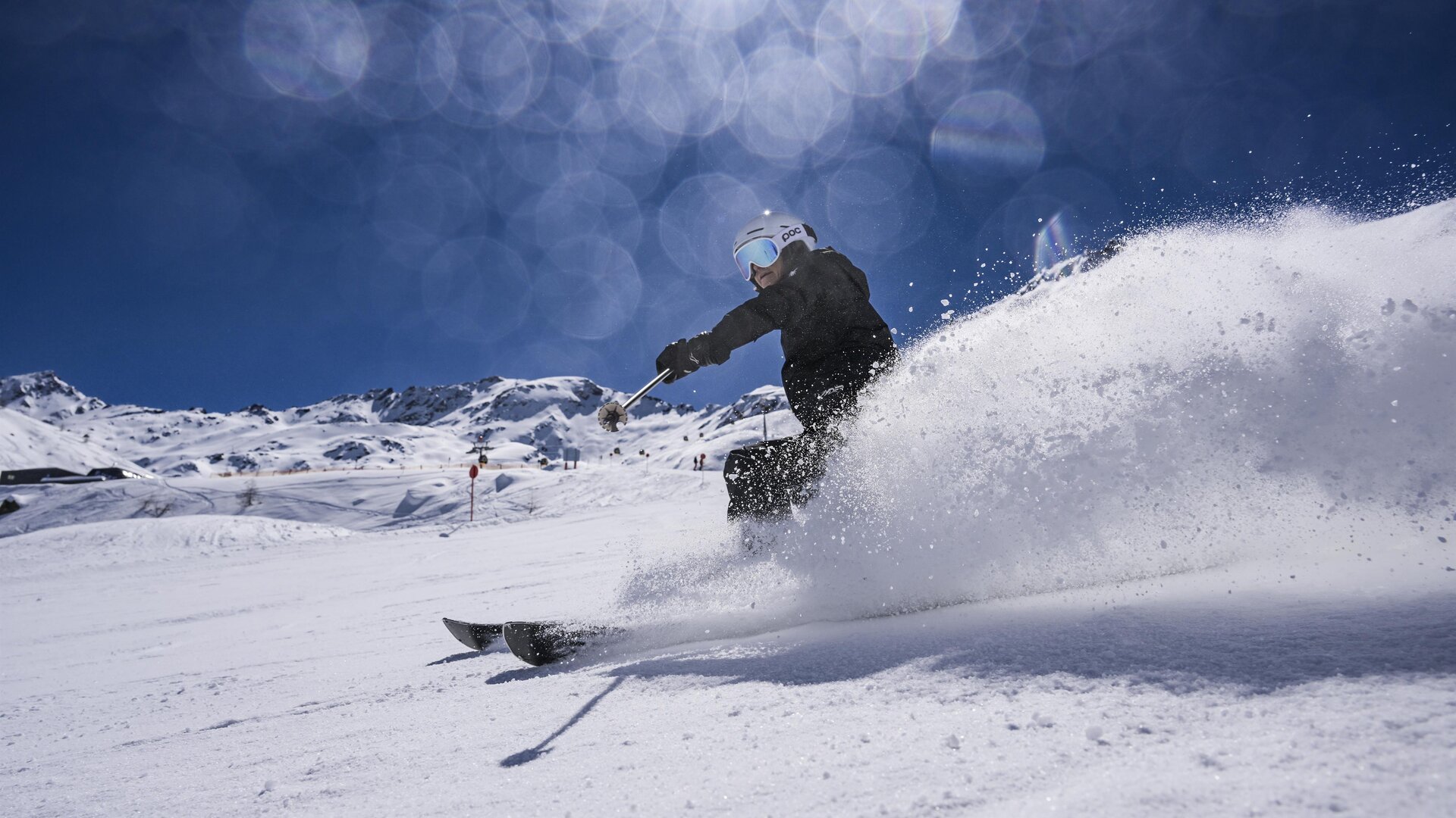 A skier glides with energy through the fresh snow. In the background, there are snow-covered mountains and a clear blue sky.