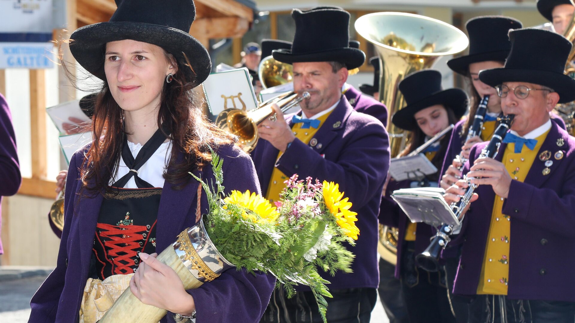 A group of people in traditional costumes is marching through the city. A woman is holding a bouquet of flowers in her hand.