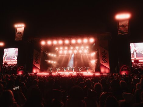 A large crowd is watching a stage performance at night. The stage is brightly lit, with red lights and several screens.
