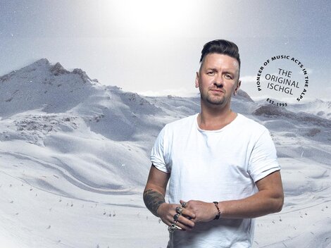 Ein Mann steht in einer schneebedeckten Berglandschaft. Die Umgebung ist weiß und frostig mit hohen Gipfeln im Hintergrund.