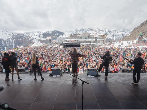 A large crowd enjoys a concert in the mountains. In the background, there are snow-covered peaks and an event building.