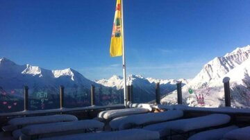 A snow-covered mountain landscape with a clear blue sky. In the foreground, deck chairs and a flag can be seen.