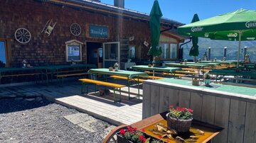 A cozy mountain cabin with green sun umbrellas and wooden tables outside. In the background, the mountains and a clear blue sky can be seen.