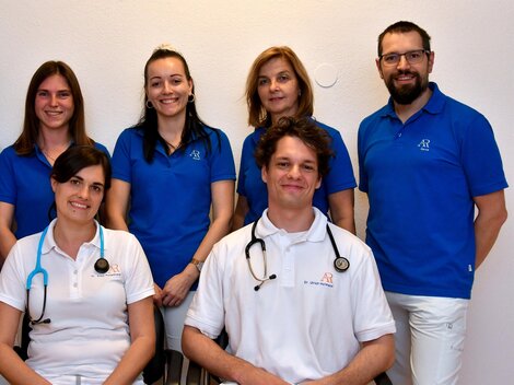 A team of professionals stands together in blue T-shirts. Two individuals are wearing stethoscopes and are smiling kindly at the camera.