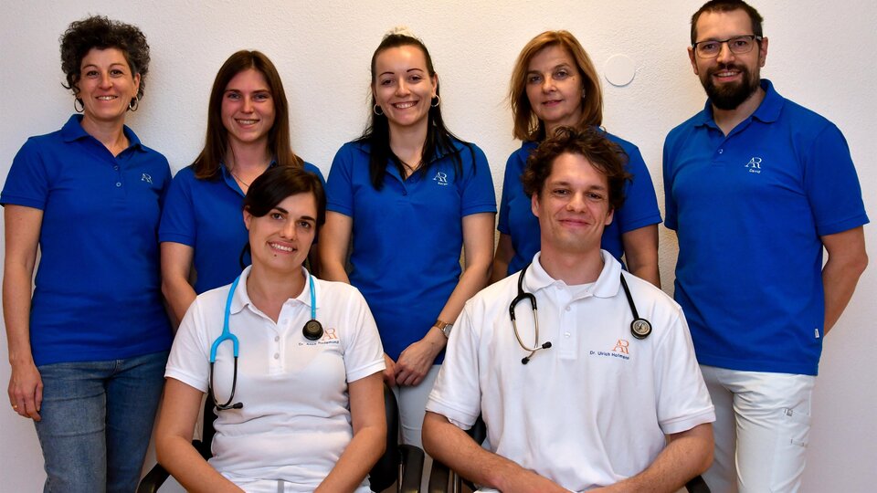 A team of professionals stands together in blue T-shirts. Two individuals are wearing stethoscopes and are smiling kindly at the camera.