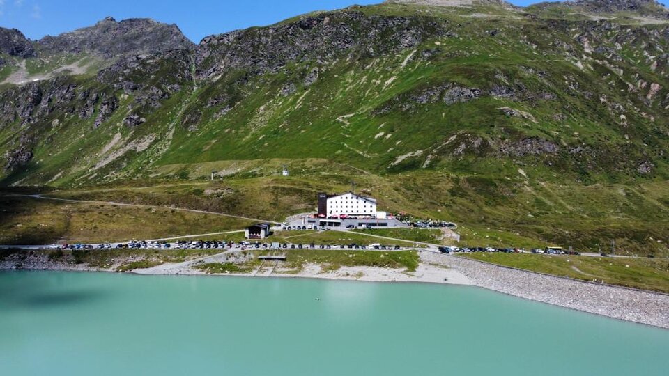 A picturesque mountain lake with bright turquoise water. A large white building stands on the shore, and green mountains surround the landscape.