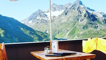 A cozy table with red seat cushions under a sun umbrella. Impressive mountains and a blue sky stretch out in the background.