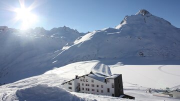 A picturesque winter landscape with snow-covered mountains. In the foreground, there is a modern hotel, surrounded by snowy slopes.