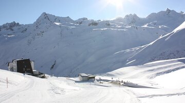A snow-covered mountain landscape with radiant sunshine. In the foreground, mountain cabins and ski slopes are visible.