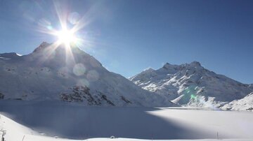 A wintry mountain landscape with snow-covered peaks and radiant sunlight. The sky is clear and blue.