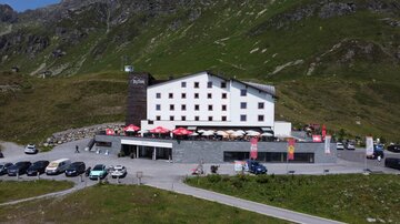A modern hotel in the mountains with a terrace and a parking lot. Surrounded by green hills and a picturesque landscape.