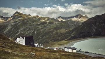 A picturesque mountain landscape with a lake in the foreground. In the background, majestic mountains and a charming accommodation can be seen.