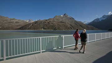 Two people are walking along a waterfront promenade overlooking a tranquil lake and majestic mountains. The sky is clear and sunny.