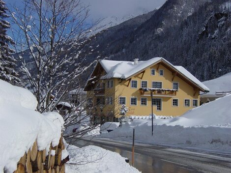 Ein charmantes, gelbes Haus im Schnee mit einem malerischen Berghintergrund. Der Weg ist von frischem Schnee bedeckt und die Bäume sind winterlich verziert.