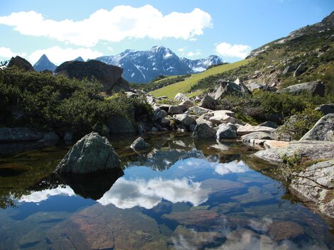 Eine malerische Berglandschaft mit hohen Gipfeln und klarem blauen Himmel. Im Vordergrund spiegelt sich das Gelände im ruhigen Wasser eines kleinen Teichs.