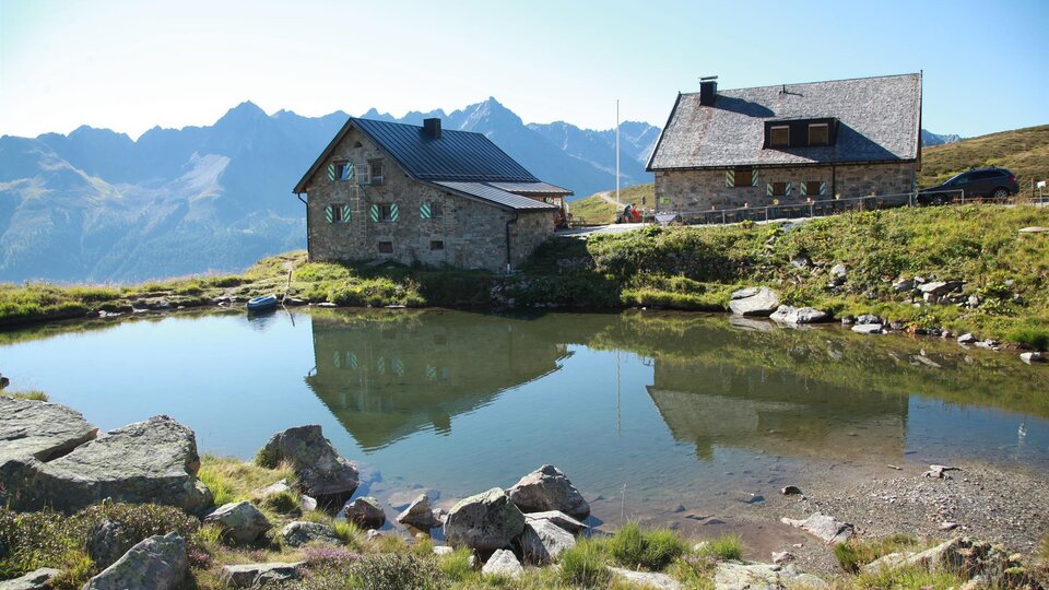 A picturesque alpine landscape with two cabins by the shore of a tranquil lake. Majestic mountains rise in the background under a clear sky.