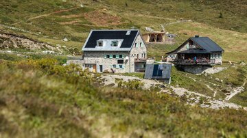 Two mountain cabins are situated in a green, hilly landscape. The surroundings are peaceful and natural.