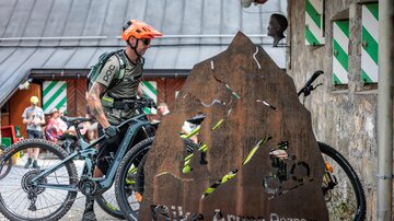A cyclist stands next to his mountain bike and a distinctive metal sign with the inscription "Bike Arena Paznaun." In the background, some buildings and a few people can be seen.
