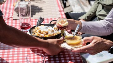 A table with a checkered tablecloth, where two people are sharing food and drinks. There is a bowl with dishes and glasses with various beverages.