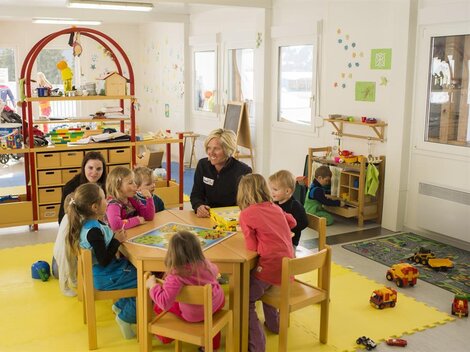 Ein heller Kindergartenraum mit Kindern, die um einen Tisch sitzen und spielen. Eine Betreuerin ist anwesend, während im Hintergrund weitere Kinder mit Spielzeug beschäftigt sind.