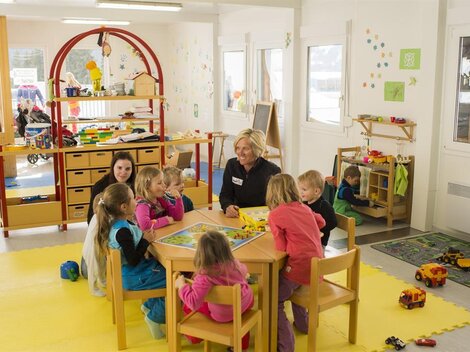 Ein heller Kindergartenraum mit Kindern, die um einen Tisch sitzen und spielen. Eine Betreuerin ist anwesend, während im Hintergrund weitere Kinder mit Spielzeug beschäftigt sind.