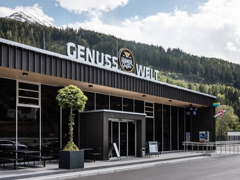 Ein modernes Gebäude mit der Aufschrift "GENUSS WELT" steht in einer grünen Landschaft. Im Hintergrund sind Berge und ein klarer Himmel zu sehen.