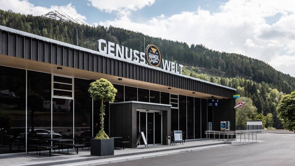 Ein modernes Gebäude mit der Aufschrift "GENUSS WELT" steht in einer grünen Landschaft. Im Hintergrund sind Berge und ein klarer Himmel zu sehen.