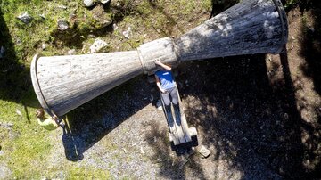 Eine große Holzskulptur in Form eines Trichters liegt im Freien. Zwei Personen interagieren mit der Skulptur auf einer grünen Wiese.