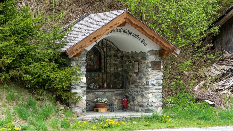 Eine kleine Kapelle aus Stein mit einem Holzdach steht in der Natur. Umgeben von Bäumen und Gras bietet sie einen ruhigen Rückzugsort.