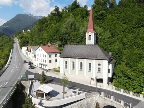 A small church with a red roof and a tall tower stands by a road. Surrounded by green hills and residential buildings.